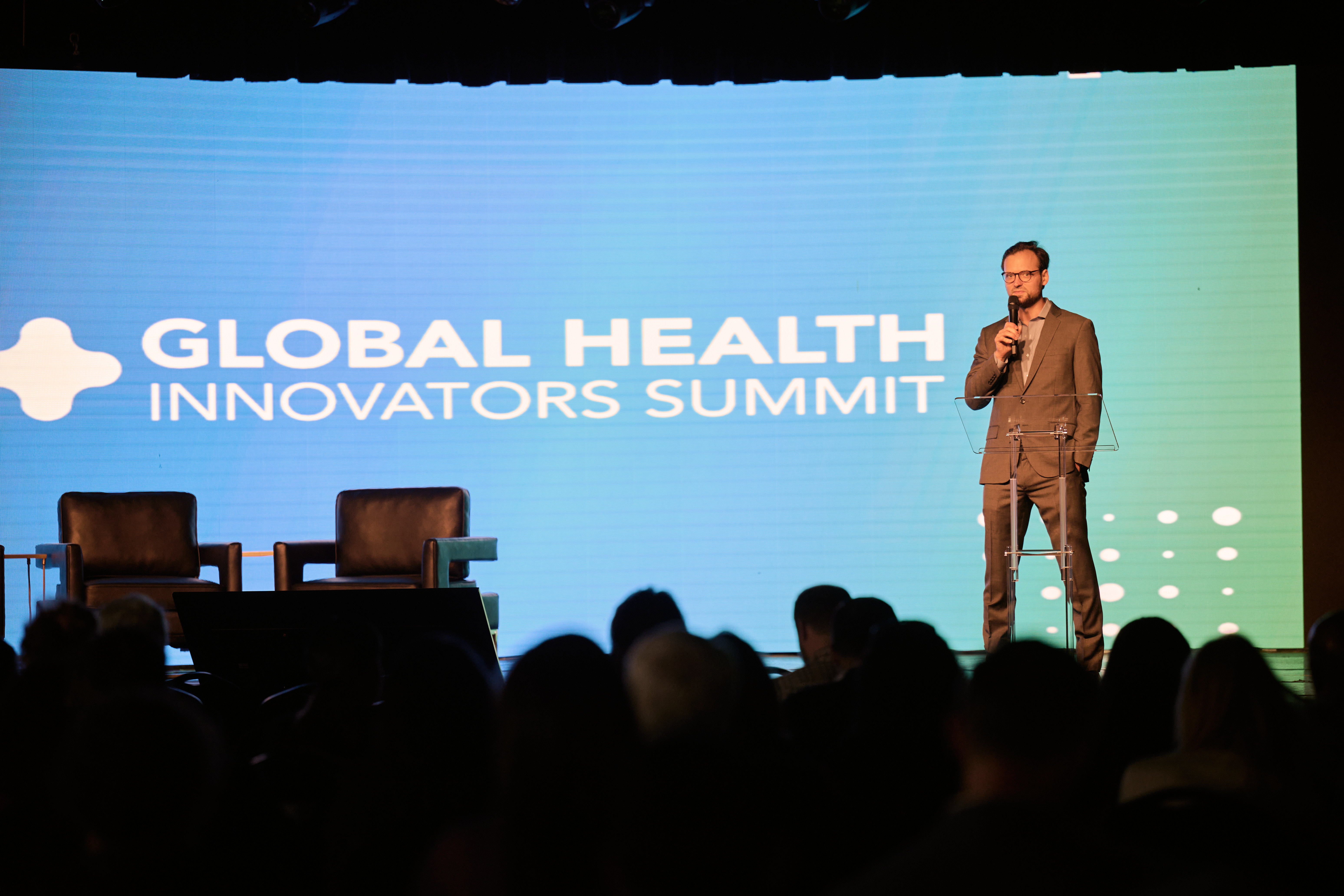 Conference presentation with man in brown suit speaking on stage with microphone, with GLOBAL HEALTH INNOVATORS SUMMIT branding visible on large screen behind him and audience silhouettes in foreground at Global Health Innovators Summit
