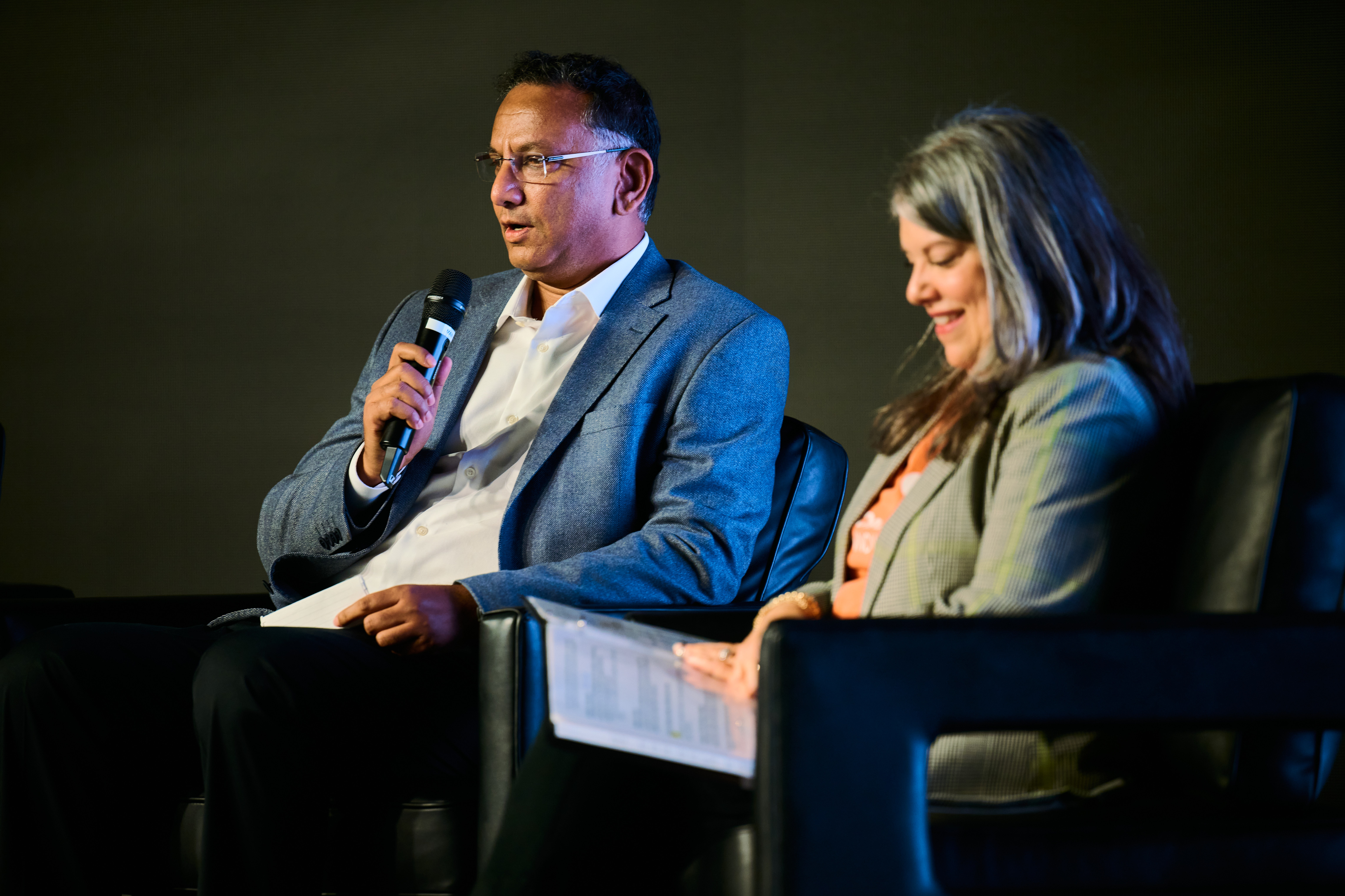 Conference speaker - man with glasses in gray suit speaking into microphone alongside woman in light blazer during Global Health Innovators Summit session