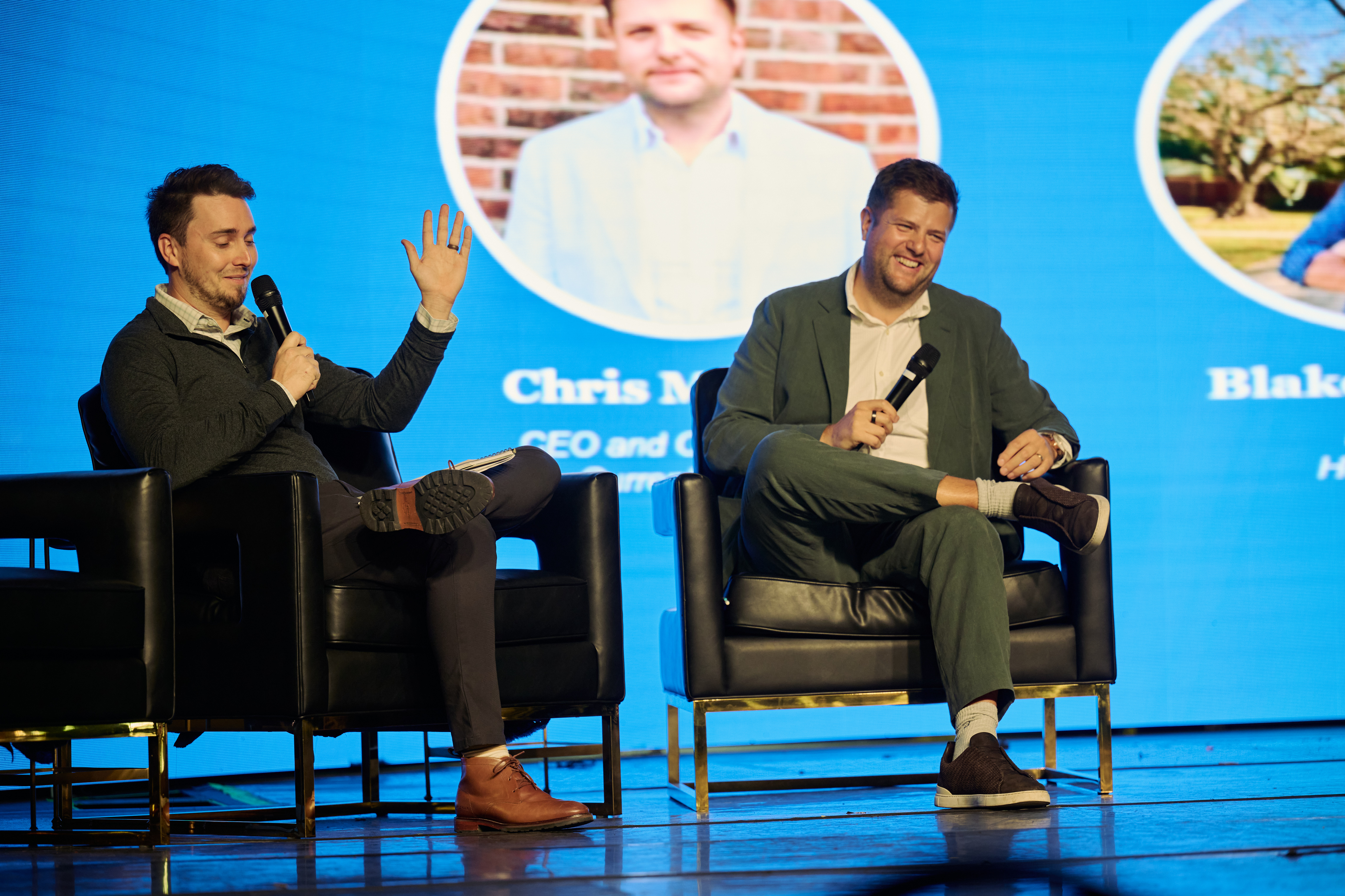 Conference discussion featuring two men seated in black chairs having a conversation with speaker photos displayed on a blue screen behind them during Global Health Innovators Summit
