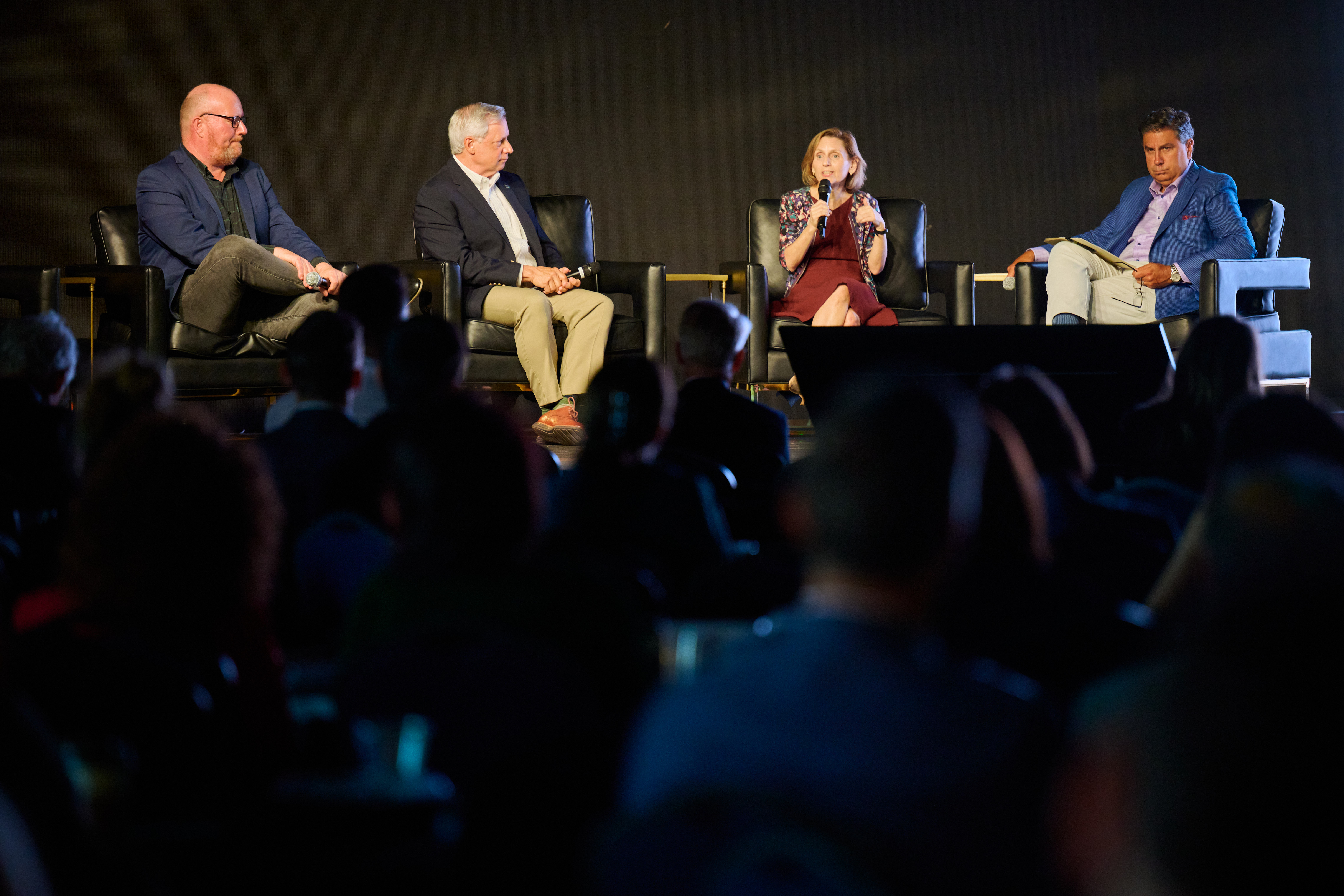 Panel discussion featuring four panelists - three men and one woman - seated on black chairs during conference session, with audience members visible in foreground at Global Health Innovators Summit