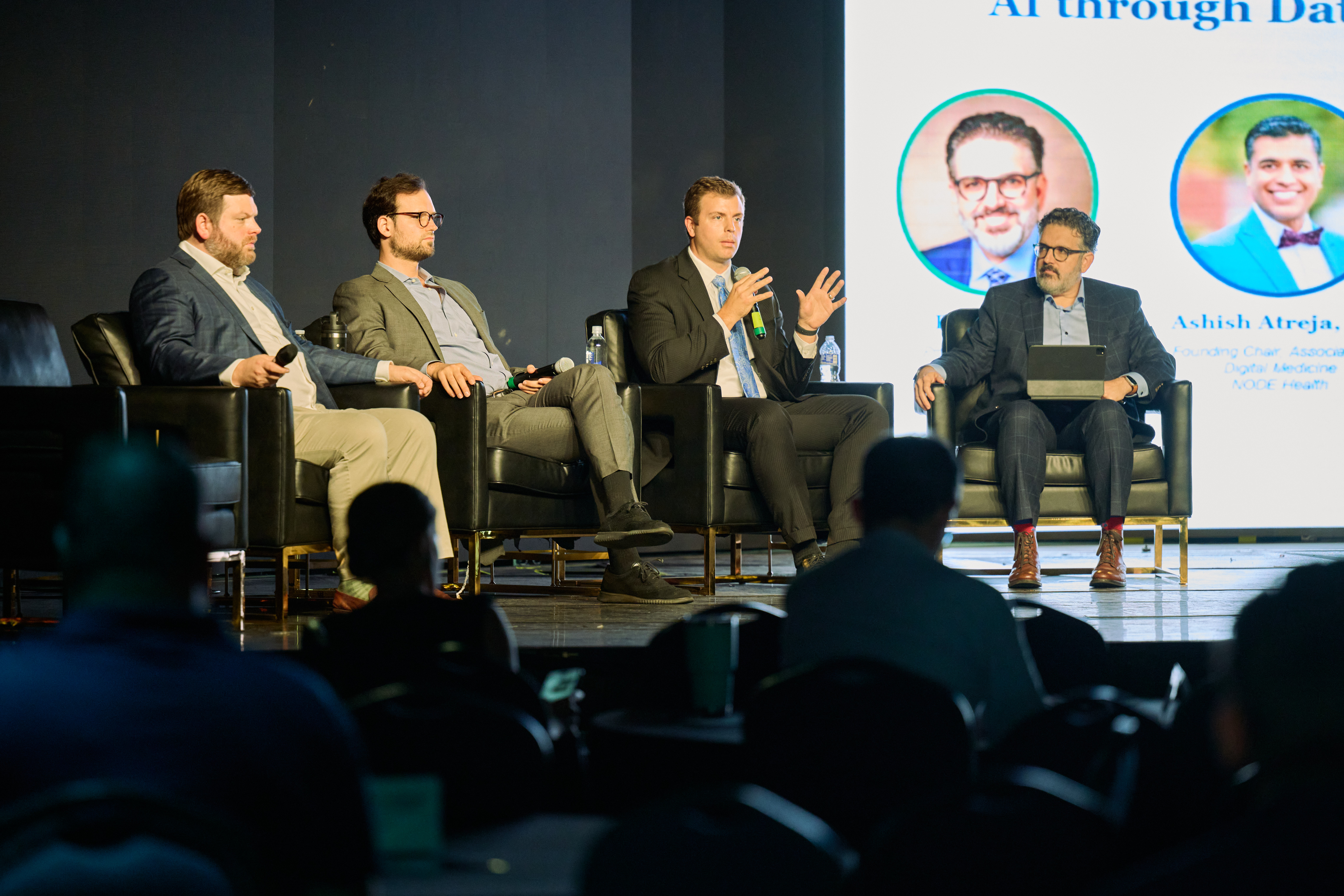 Panel discussion featuring four men seated on black chairs during conference session with audience members visible in foreground and speaker photos displayed on screen behind them at Global Health Innovators Summit