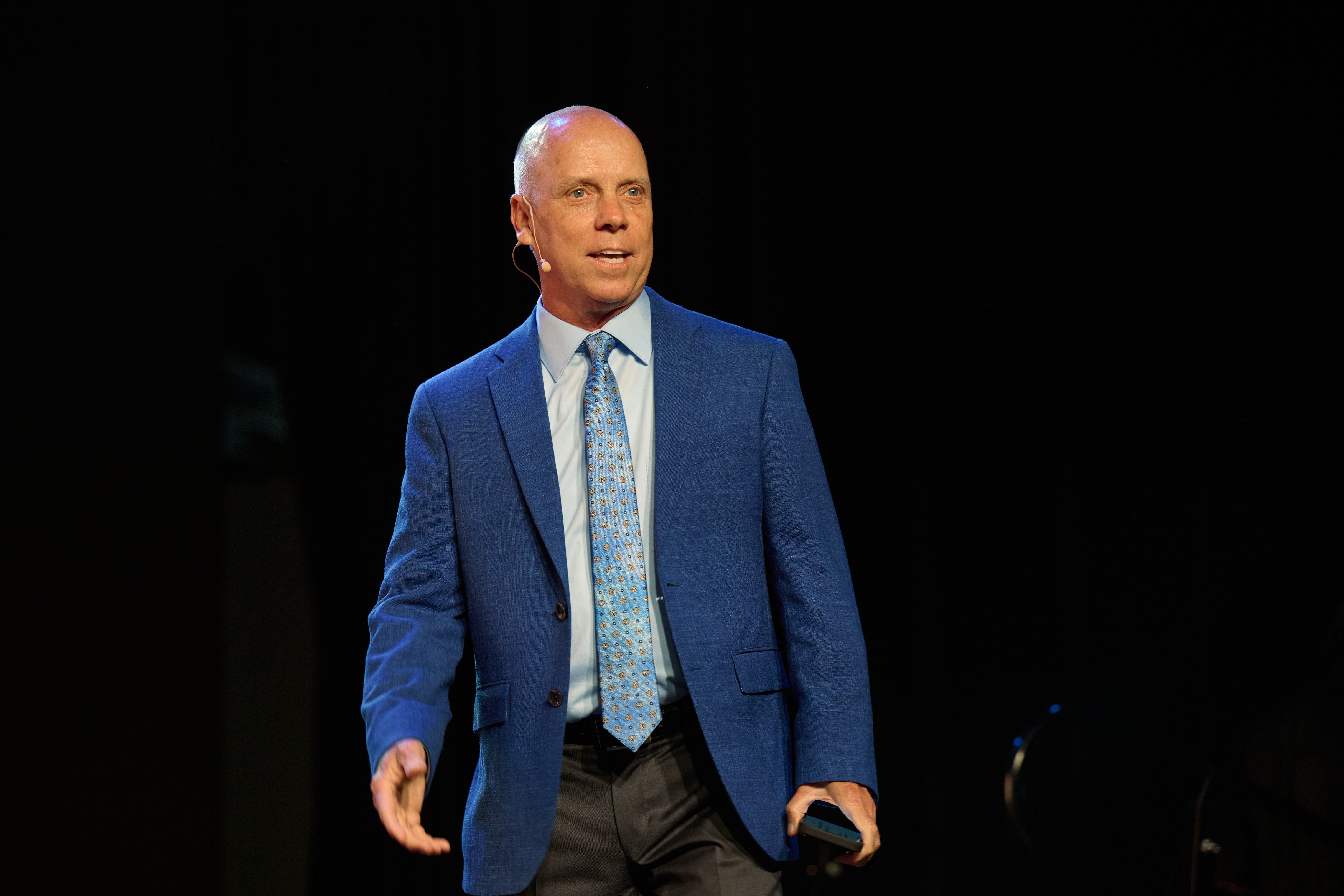 Keynote speaker - man in blue suit with microphone speaking on stage against dark background at Global Health Innovators Summit