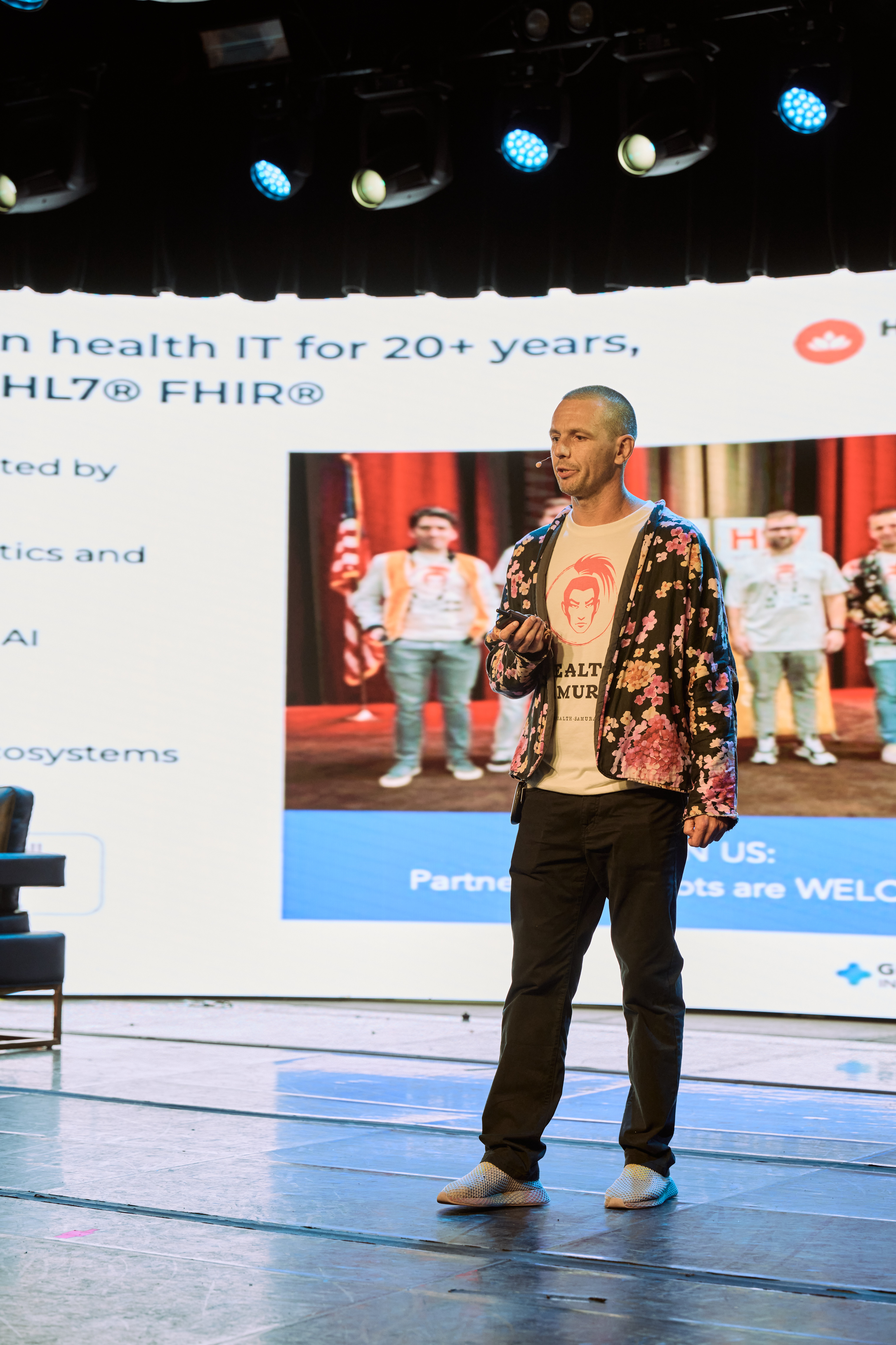 Keynote speaker - man in floral jacket presenting on stage at Global Health Innovators Summit with audience and conference branding visible