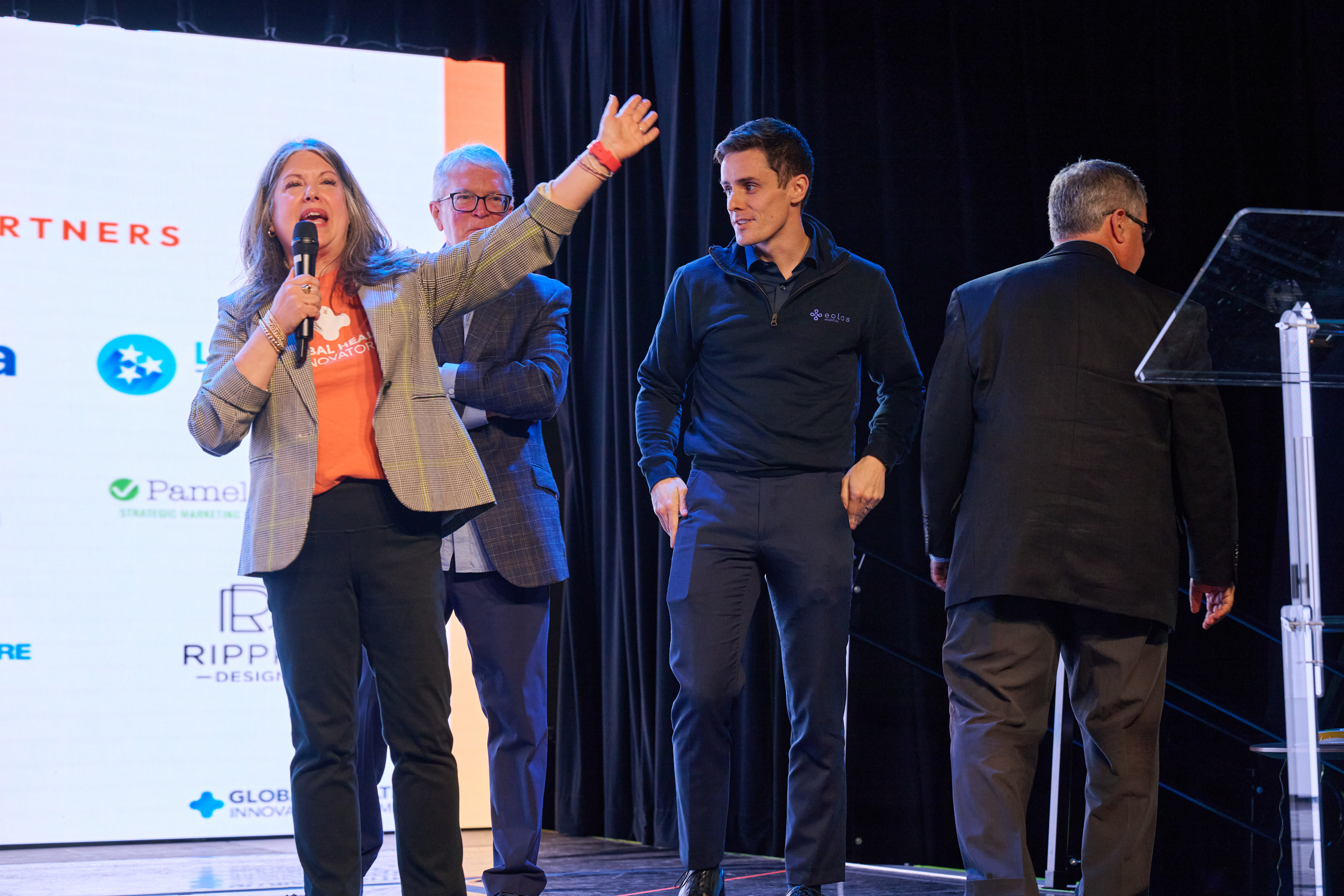 Award presentation ceremony showing people on stage during what appears to be an awards ceremony with sponsor logos visible in the background, including a woman speaking into a microphone at Global Health Innovators Summit