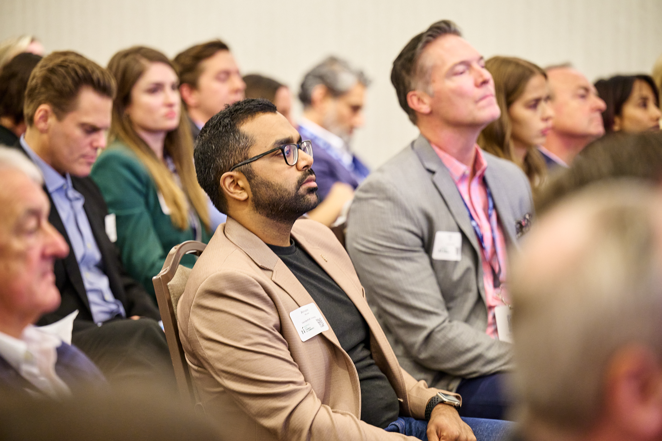 Conference audience photo showing engaged attendees listening attentively to a presentation. In the foreground, a man with glasses and a beard wearing a tan/beige blazer over a dark shirt is prominently featured, along with a man in a gray suit with a pink lanyard. The audience includes diverse professionals in business attire, with a woman in a teal/green blazer visible in the background. The setting captures the focused, professional atmosphere of the Global Health Innovators Summit.