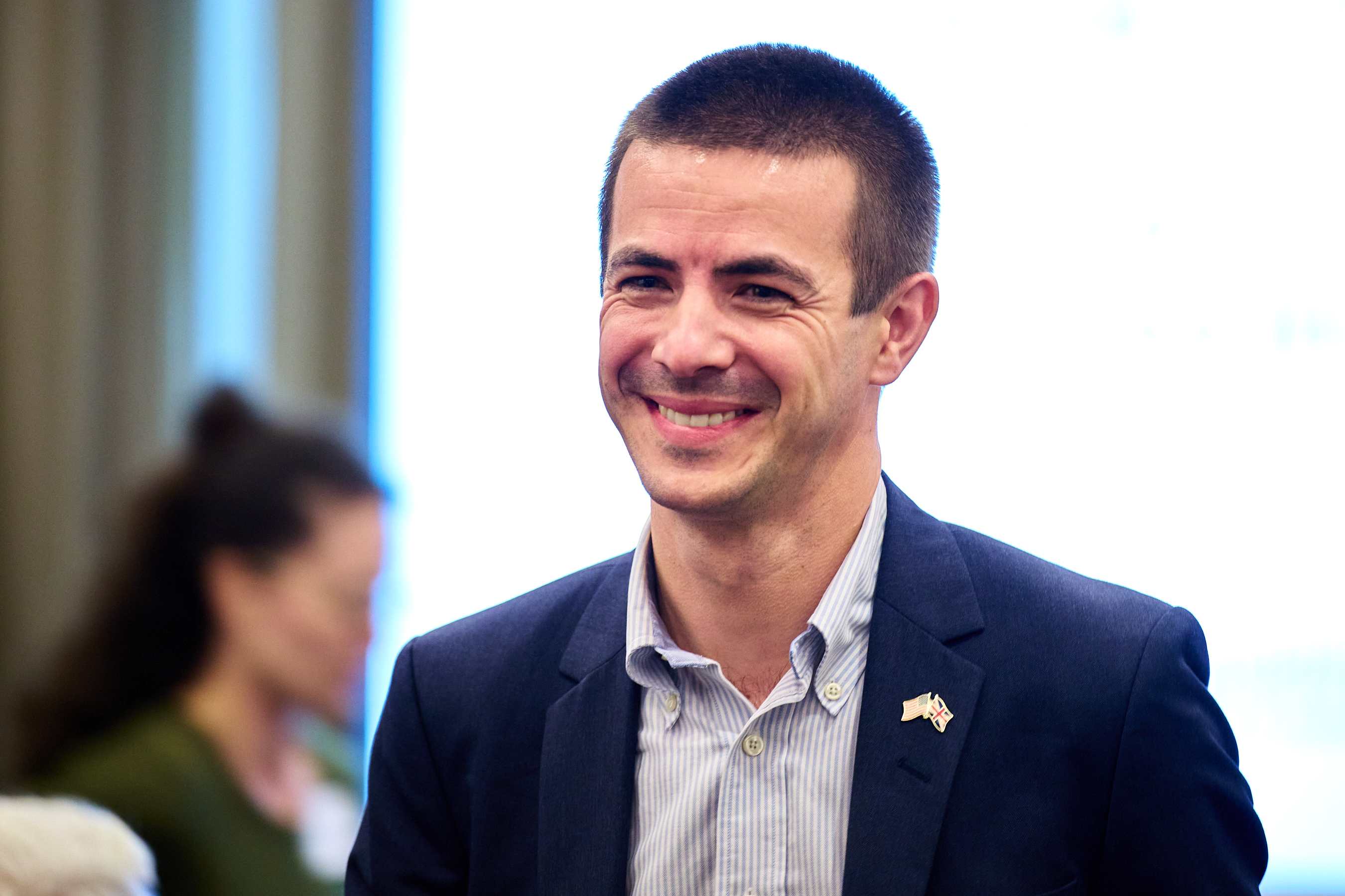Conference photo showing a professional man with short brown hair and a genuine, warm smile, wearing a navy blue business suit with a light gray and white striped dress shirt. He has a small American flag pin on his lapel and is photographed in what appears to be a bright conference venue with natural lighting from windows in the background. The man appears confident and approachable, typical of a healthcare industry professional or conference attendee.