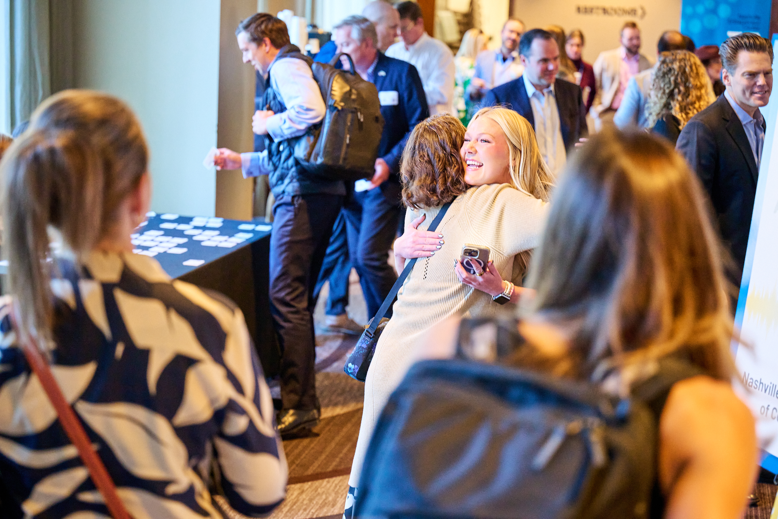 Conference networking photo showing a warm moment between two women embracing in what appears to be a conference hallway or networking area. One woman has blonde hair and is wearing a light-colored sweater, hugging another woman from behind. The background shows other conference attendees in business attire mingling, with elegant venue lighting and a professional conference atmosphere. The scene captures the personal connections and networking spirit of the health innovation summit.