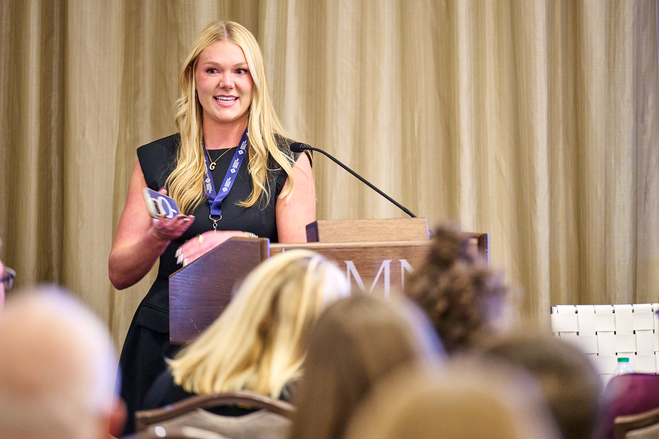 Conference presentation photo showing a blonde woman in black sleeveless top with blue lanyard speaking at wooden podium with microphone. She's holding what appears to be an award or recognition item, addressing an audience with attendees visible from behind in the foreground. The professional conference setting shows elegant curtains and proper staging at the OMNI venue.