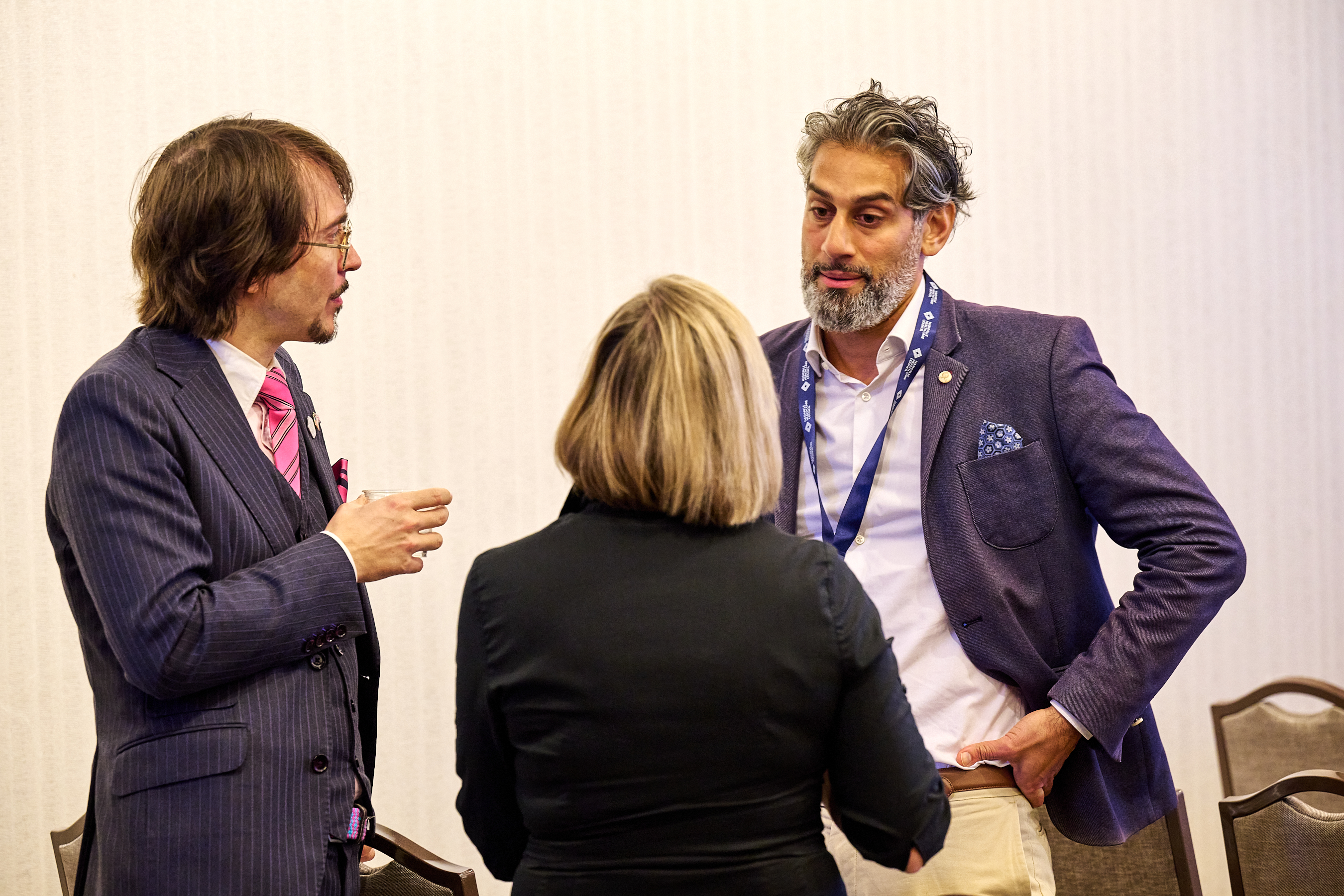 Conference networking conversation photo showing three professionals engaged in discussion - a man in pinstripe suit with pink tie on the left, woman with blonde bob in black blazer in the middle (from behind), and man with salt-and-pepper hair in purple blazer with blue lanyard on the right.