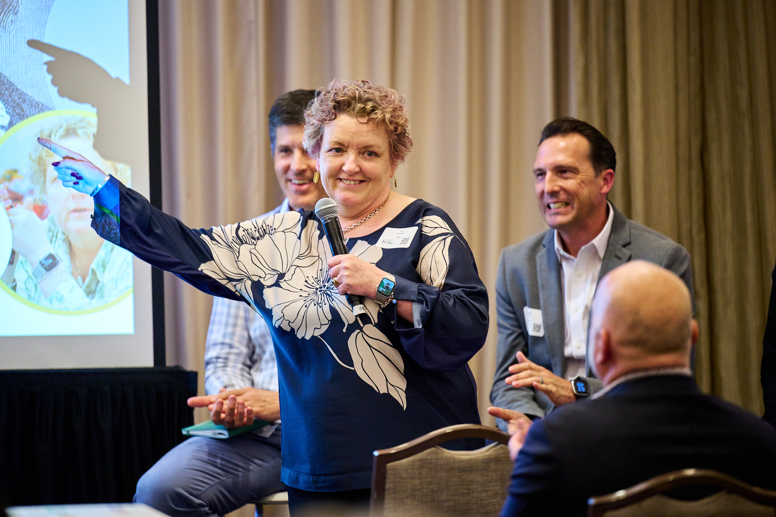 Conference presentation photo showing a woman with curly hair wearing a navy blue top with large white floral print, holding a microphone and speaking animatedly at a conference session. She appears to be presenting or facilitating a discussion, with other conference attendees visible in the background and a presentation screen showing what appears to be a map or data visualization. The setting captures the engaging, interactive nature of the health innovation conference.