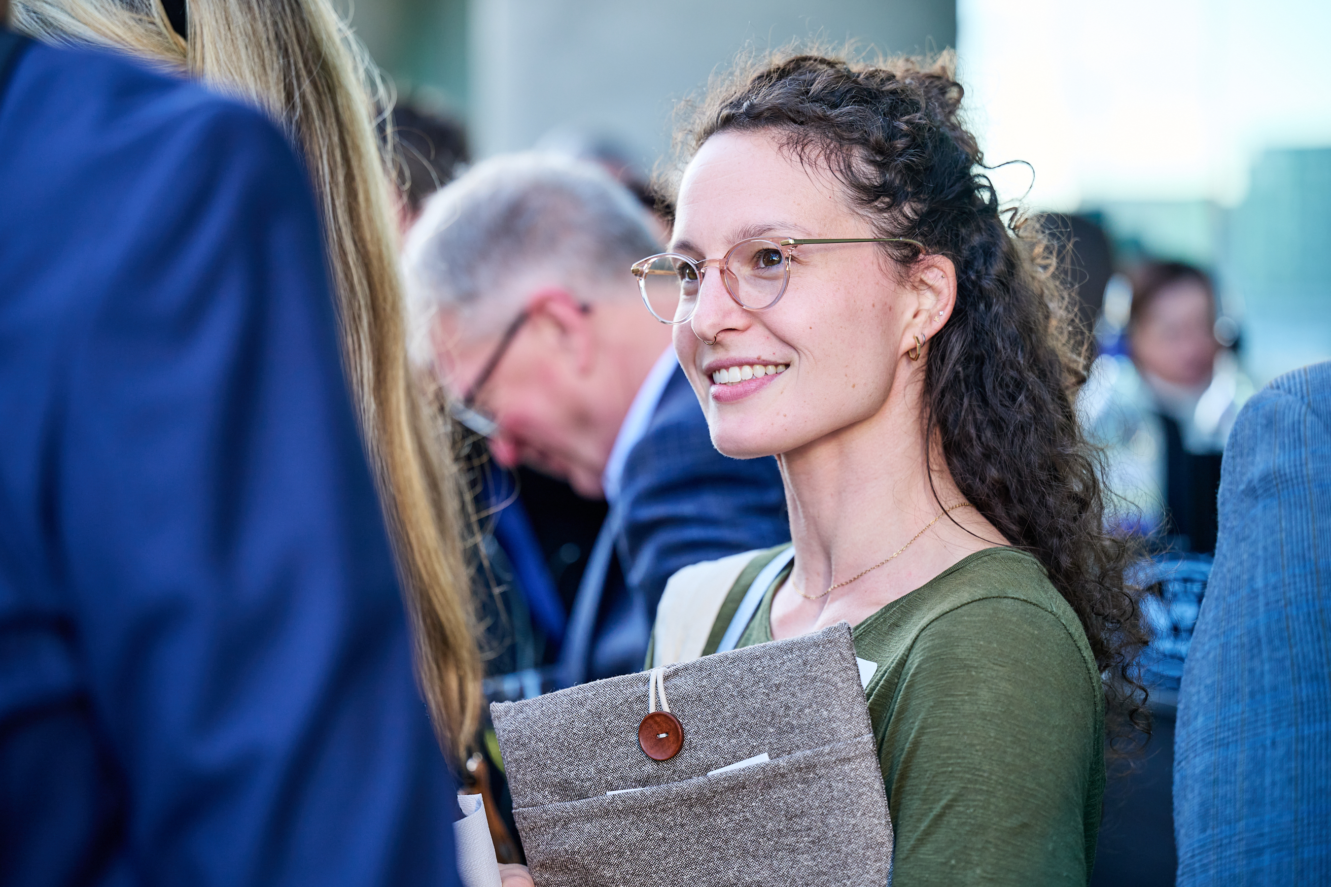Conference networking photo showing a young woman with curly dark hair, glasses, and a warm smile wearing a green top and holding a gray laptop bag with brown button closure. She's at a networking event with other conference attendees visible in the background, creating a candid, professional networking atmosphere typical of health innovation conferences.
