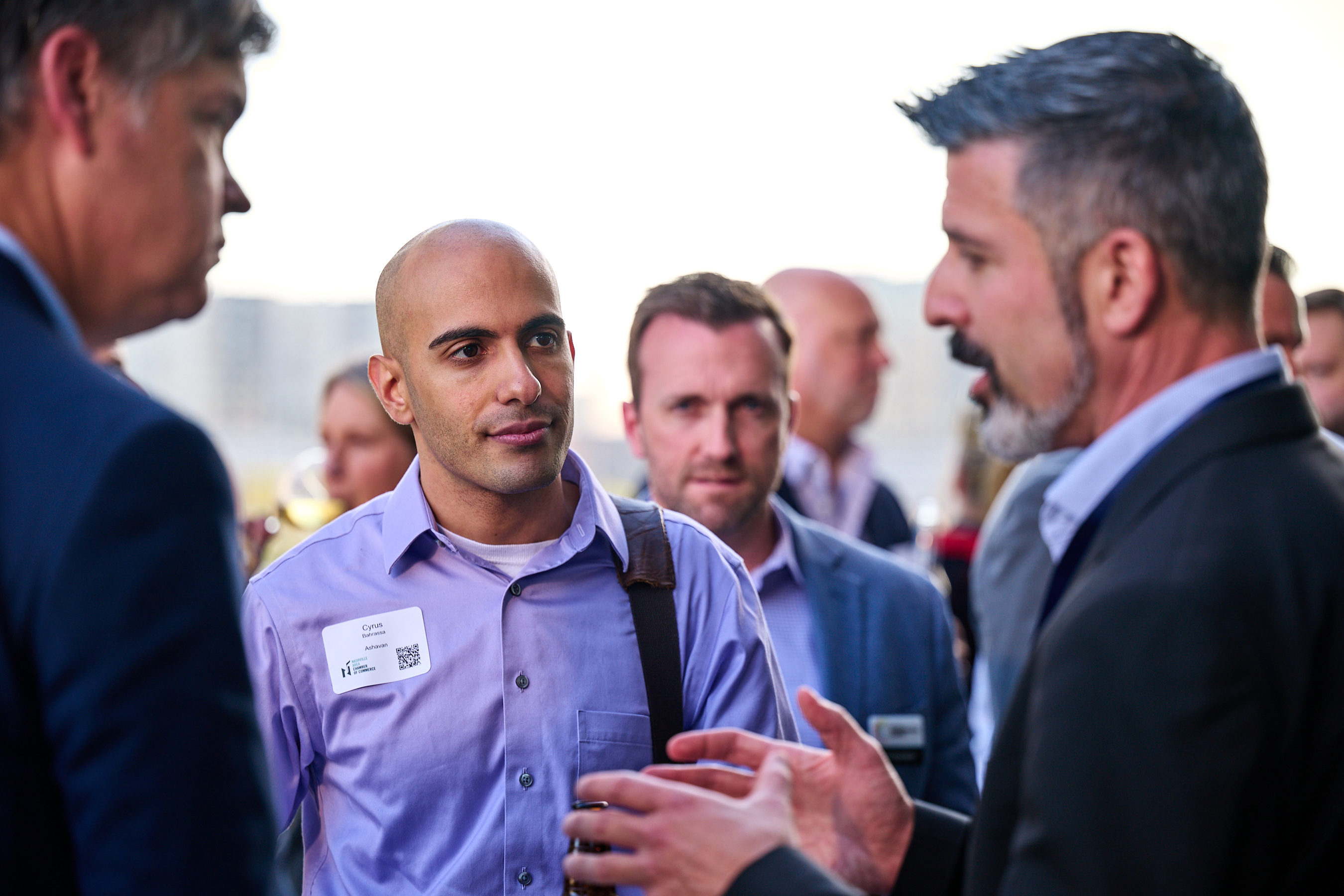 Conference networking photo showing a bald man in the center wearing a light purple/lavender button-down shirt with a white name tag, engaged in conversation with other professionals. To his left is a man in a navy suit, and to his right is another man with dark hair and beard. The background shows other conference attendees networking in what appears to be a bright conference venue with large windows. The scene captures the engaged, professional networking atmosphere of the health innovation summit.