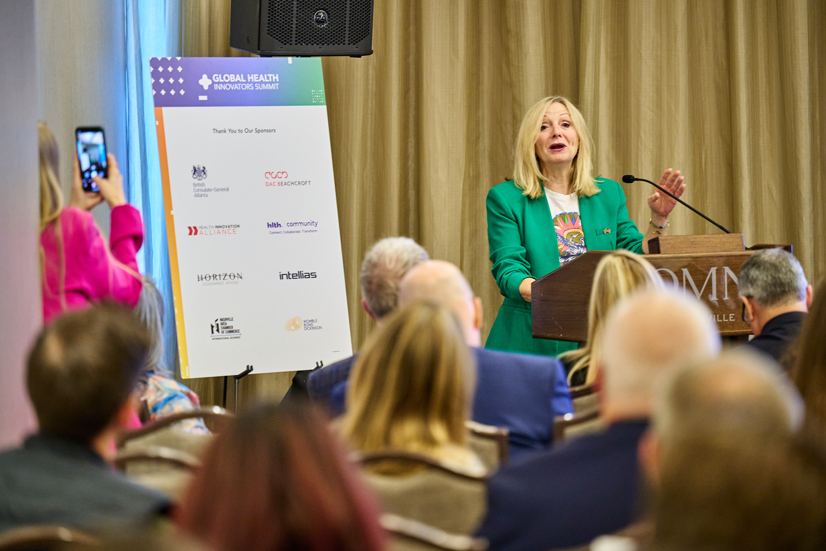 West Yorkshire Mayor GHIS Conference - Blonde woman in bright green blazer speaking enthusiastically at wooden podium with microphone, addressing full audience with sponsor banners including 'Global Health Innovators Summit' and 'Thank You to Our Sponsors' visible at OMNI Nashville venue