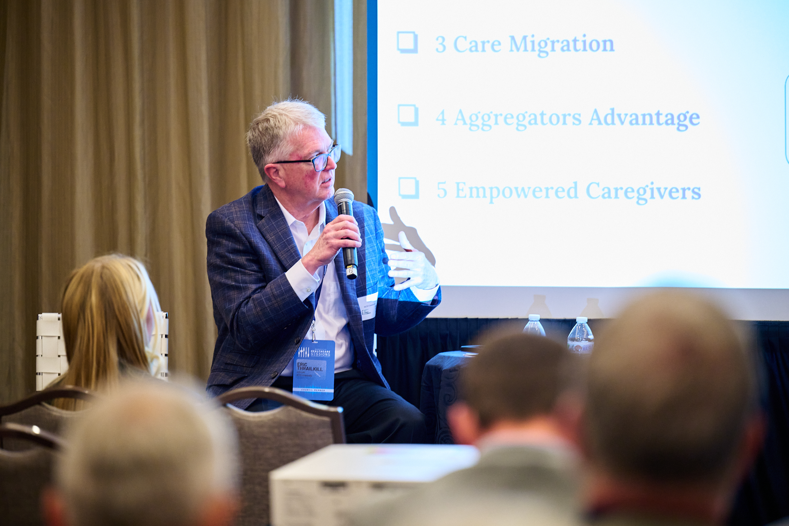 Conference presentation photo showing a man with gray hair and glasses wearing a dark plaid suit jacket with white shirt and blue conference lanyard, speaking into a microphone while moderating a session. In the background is a large presentation screen displaying '3 Care Migration', '4 Aggregators Advantage', and '5 Empowered Caregivers' with blue checkmarks. Audience members with blonde hair are visible from behind in the foreground, and the setting shows elegant curtains and professional conference staging, capturing the educational aspect of the health innovation summit.