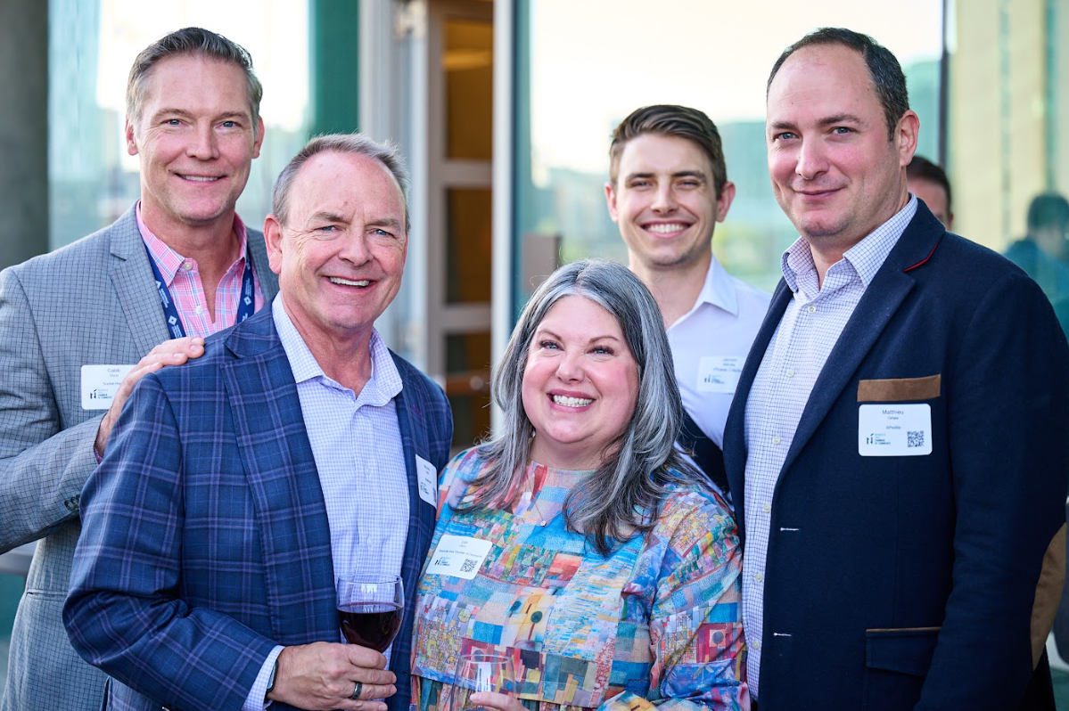 Conference group networking photo showing five professionals in a friendly group pose at a health innovation conference. From left to right: a man in gray suit with pink shirt and blue lanyard, a man in blue plaid blazer holding wine glass, a woman in center wearing colorful patterned top, a young man in light blue shirt, and a man in navy blazer with checkered shirt. All are smiling warmly in a bright conference networking area with natural lighting, capturing the collaborative spirit of the health innovation summit.